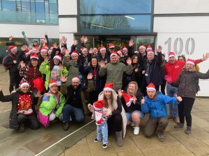 A group of more than 30 people, some in xmas hats, standing in a group and waving at the camera