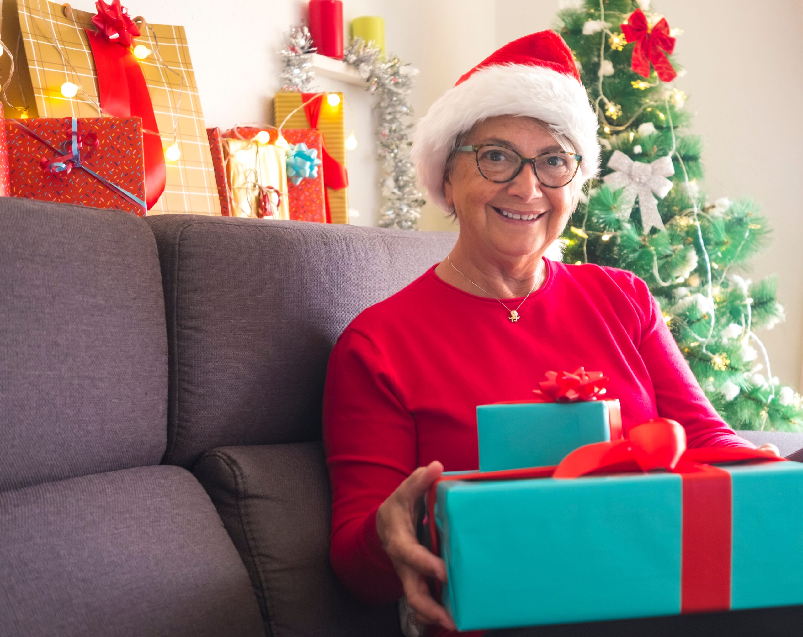 Smiling older adult in a Santa hat holding wrapped gifts beside a decorated Christmas tree.